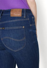 Vaqueros de mezclilla azul oscuro con textura suave, bolsillos traseros curvados y una etiqueta de cuero marrón en la cintura con el nombre de la marca "Lee".