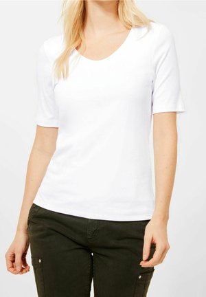Frau trägt ein einfaches weißes T-Shirt mit kurzen Ärmeln und dunkelgrüne Hosen mit Seitentaschen, steht vor hellem Hintergrund.