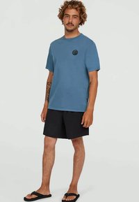 Man with curly hair wearing a blue logo T-shirt, black shorts, flip-flops, standing against a neutral background.
