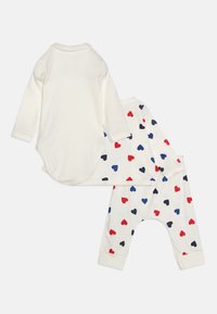 Hvitt langermet bodysuit og mønstrede bukser med røde og blå hjerter på kremfarget stoff. Mykt bomullsmateriale, uformell design.