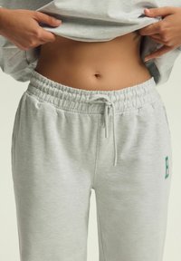 Jasnoszare dresy wykonane z miękkiego materiału, z elastycznym pasem w talii z sznurkiem oraz małym zielonym logo w kształcie litery na lewej stronie.