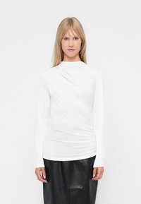 Lange mouw witte top met een gedrapeerd ontwerp en textuurstof. Het shirt heeft een hoge halslijn en valt boven een zwarte rok.