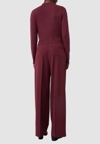 Bordeaux-farbenes Longsleeve-Oberteil mit tailliertem Schnitt und elastischem Bund, kombiniert mit hochgeschnittenen, weit geschnittenen Hosen in der gleichen Farbe. Glatte Textur.