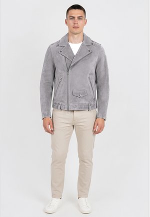 Muškarac okrenut prema naprijed nosi sivu biker jaknu od brušene kože, bež hlače, bijele tenisice i bijelu majicu na jednobijeloj pozadini.