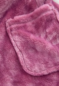 Material moale și plușat de culoare roz, cu o textură pufoasă; are un buzunar mic cusut. Designul general este confortabil și tactil.