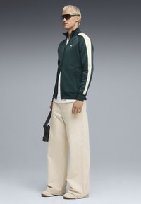 Puma Trackjacke in Dunkelgrün mit cremefarbenen Streifen, mit Reißverschluss, kombiniert mit lockeren cremefarbenen Hosen und einer dunklen Tasche. Das Modell trägt Sonnenbrillen.