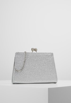 Pochette à paillettes argentées avec une petite chaîne métallique et un fermoir boule rond, posée sur une surface blanche avec un fond gris clair.