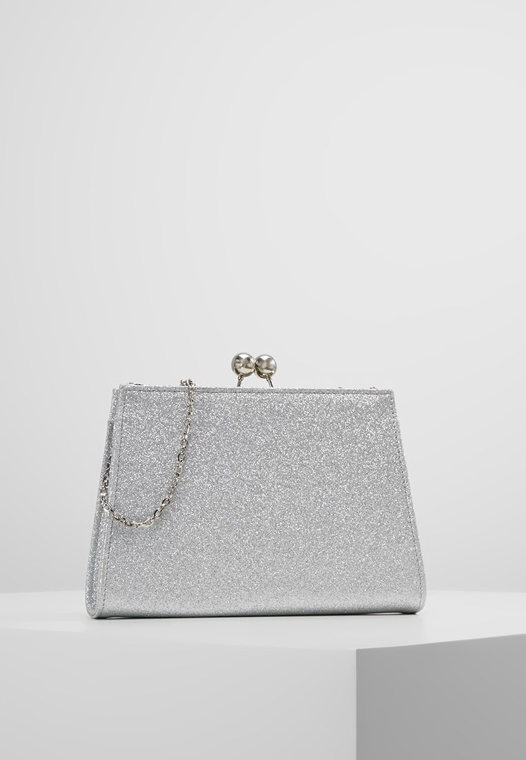 Pochette à paillettes argentées avec une petite chaîne métallique et un fermoir boule rond, posée sur une surface blanche avec un fond gris clair.