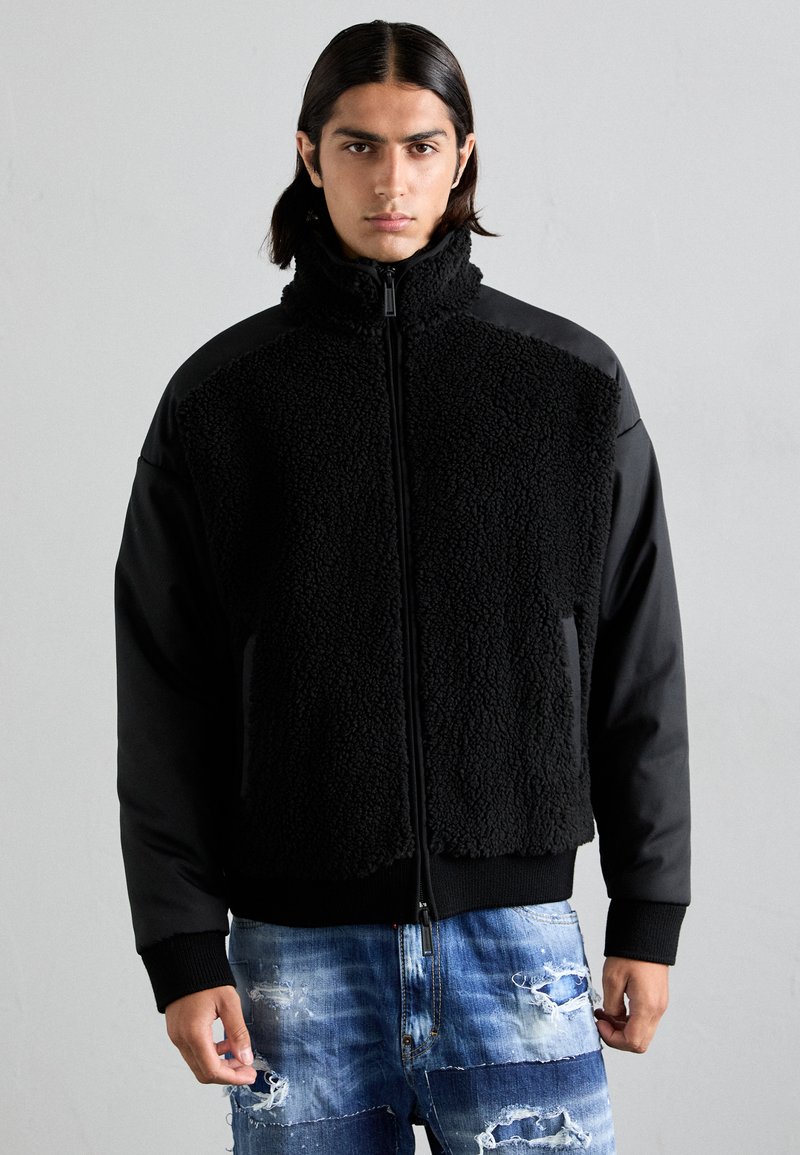 Schwarze Jacke mit strukturiertem Fleece-Front, glatten Ärmeln, hohem Kragen und Reißverschluss. Tragen mit zerrissenen blauen Jeans.