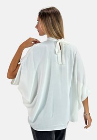 Blouse blanche ample à manches chauve-souris, avec un lien au niveau de la nuque à l'arrière, en tissu doux et texturé.