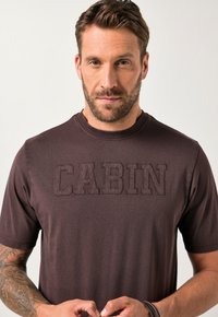 Mørkebrun t-shirt med rund hals og tekstureret "CABIN"-bogstaver over brystet. Korte ærmer, afslappet pasform og blødt stof.