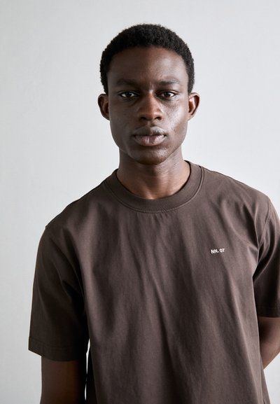 T-shirt en coton marron à manches courtes et col rond, avec un petit logo blanc sur la poitrine. Texture lisse et coupe décontractée.