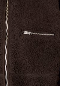 Brązowa polarowa kurtka o teksturowanej powierzchni, z metalowym zamkiem błyskawicznym w kieszeni oraz dopasowanym zamkiem z przodu.