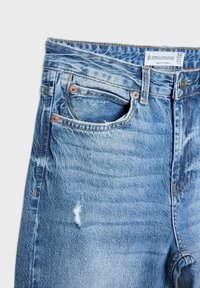 Primer plano de jeans de mezclilla azul que muestra el bolsillo delantero, pequeño desgaste, remaches de cobre, cierre de botón y etiqueta de la marca Stradivarius con talla 38.