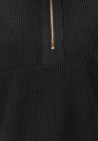 Schwarzer gerippter Pullover mit hohem Kragen und goldfarbenem Reißverschluss. Texturiertes Gewebe mit vertikalen Linien. Verfügt über dreieckige Schulterdetails.