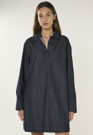 Kobieta o długich brązowych włosach nosi luźną, ciemną sukienkę koszulową z długim rękawem i kołnierzykiem, stojąc na jednolitym tle.