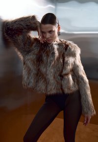 Chaqueta corta de pelo sintético en tonos marrones con una textura esponjosa y mangas anchas, combinada con un bodysuit negro y grandes pendientes geométricos.