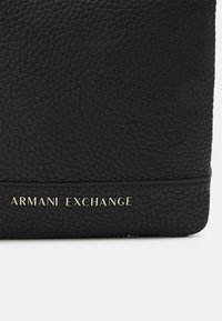 Crna kožna torba s teksturiranim završetkom, sadrži horizontalnu crtu i urezano ime branda "ARMANI EXCHANGE" u srebrnoj boji.