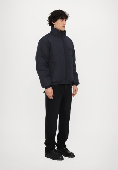 Veste rembourrée bleu marine avec col haut et fermeture éclair sur le devant, dotée de poignets et d'un ourlet élastiques. Portée avec un pantalon noir et des chaussures noires.
