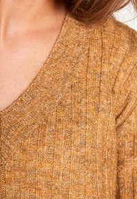 Stickad tröja med djup V-hals, ribbad struktur, i en varm senapsfärg. Har ett mjukt, medeltungt tyg med subtila inslag.