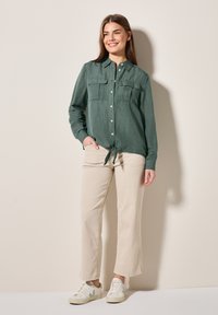 Camicia verde con bottoni, due tasche sul petto e fondo annodato. Abbinata a pantaloni beige a gamba larga e sneakers bianche. Tessuto morbido.