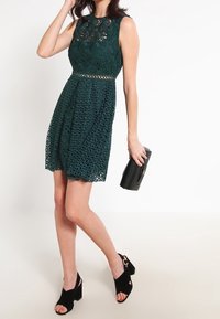 Lace cocktailklänning i mörkgrön, ärmlös med en figursydd överdel, utsvängd kjol, invecklade blommönster och en midjeförskärning. Svart kuvertväska och klackar.