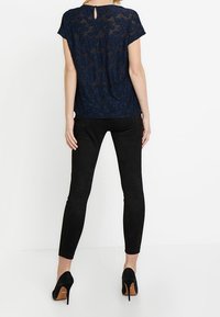 Granatowa bluzka z koronką, z krótkimi rękawami i wycięciem na plecach, w połączeniu z czarnymi dopasowanymi spodniami oraz czarnymi szpilkami.