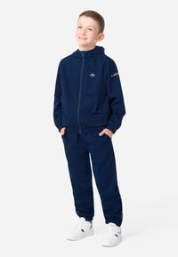 Tmavomodrá tepláková súprava z ľahkej látky, s mikinou na zips a elastickými nohavicami. Logo Lacoste na hrudi a rukáve.
