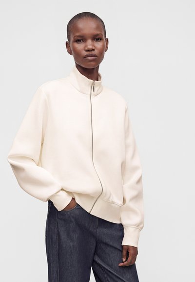 Personne portant un sweat-shirt crème à col montant zippé avec manches longues et pantalon sombre, debout avec une main dans la poche.