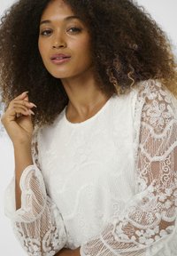 Blouse blanche avec une broderie florale élaborée, des manches en dentelle transparente et un col rond. La texture douce et les motifs délicats sont mis en avant.