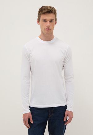 Joven mirando hacia adelante, vistiendo una camiseta blanca de manga larga lisa y jeans azul oscuro, sobre un fondo neutro.