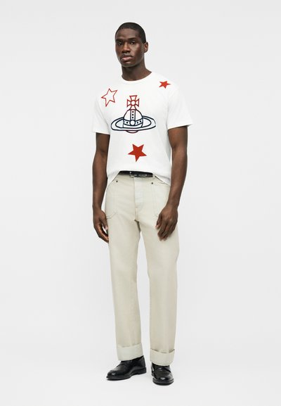 Άνδρας που φοράει λευκό μπλουζάκι με κόκκινα αστέρια και έμβλημα, μπεζ παντελόνι με μανσέτες, μαύρα παπούτσια, στέκεται μπροστά σε απλό φωτεινό φόντο.