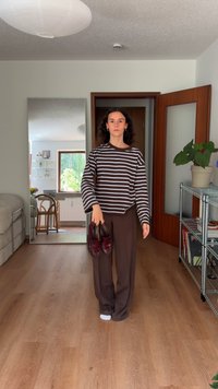 Brown and white striped long-sleeve shirt, dark brown wide-leg pants, holding burgundy shoes, standing in a room with light wooden floor.