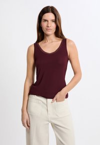 Canotta senza maniche color bordeaux con scollo a V e rifiniture texturizzate, realizzata in tessuto morbido, abbinata a pantaloni chiari e larghi.