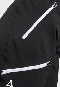 Schwarze Jacke mit mehreren weißen Reißverschlusstaschen. Das Material hat eine glatte, strukturierte Oberfläche mit sichtbaren Nähten und einem Logo-Akzent.