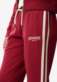 Red sweatpants featuring white side stripes and an embroidered "SUPERDRY ATHLETICS" logo. Made of soft material with a relaxed fit.