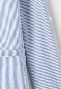 Tessuto a righe blu e bianche, caratterizzato da linee orizzontali e verticali, con colletto, dettaglio con bottoni sulla manica e accenti cuciti.