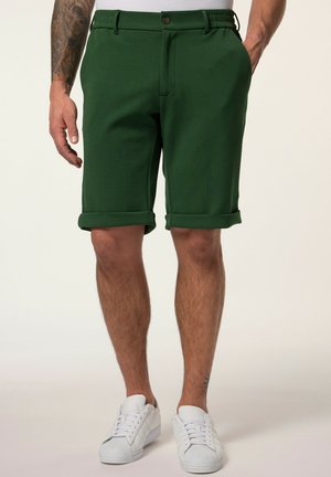 Mand iført mørkegrønne shorts med opsmøgede kanter, hvide sneakers, med tatoveret venstre arm og nederste højre ben synligt mod ensfarvet baggrund.