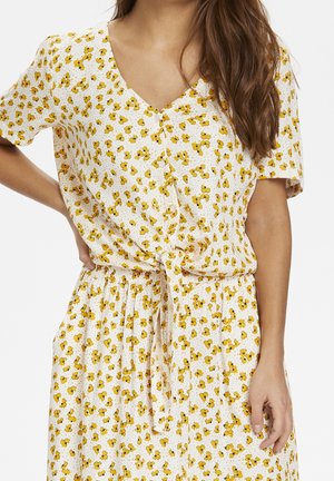 Femme portant une robe blanche à manches courtes avec un motif floral jaune, nouée à la taille, debout avec une main sur la hanche.