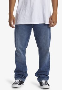 Vaqueros de mezclilla azul con un corte recto, con un lavado desgastado y ligeras arrugas. Combinados con zapatillas negras y una camiseta blanca.