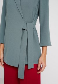 Top v tyrkysové barvě s 3/4 rukávy a zavazovacím detailem v pase, vyrobený z texturované látky. Kombinován s červenými kalhotami, což vytváří sladěný outfit.