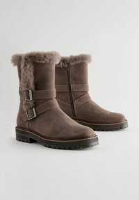 Brown suede boots featuring a faux fur collar, two adjustable buckle straps, a side zipper, and a textured rubber sole.