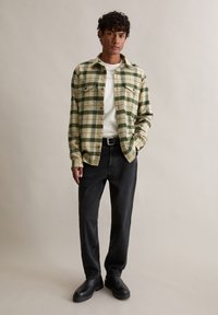 Groen- en crèmekleurig geruit flanellen overhemd met twee borstzakken, gedragen over een wit t-shirt, gecombineerd met zwarte jeans en zwarte laarzen.