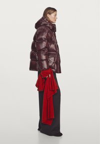 Bordeauxrode gewatteerde jas met een hoge kraag en een glanzende afwerking, gecombineerd met een rode sjaal en losse zwarte broek.