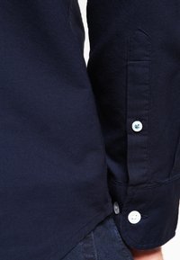 Marinblå skjorta med en texturerad väv, spetsig krage och knäppta manschetter med vita knappar och synliga stygn längs sömmen.