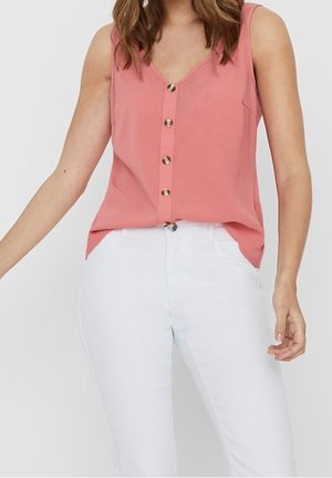 Kobieta ubrana w różową, bezrękawnikową bluzkę zapinaną na guziki, wsuniętą w białe, wysokiego stanu spodnie na jednolitym tle.