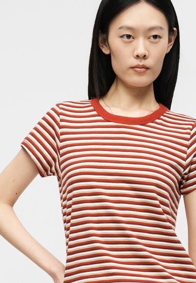 Νεαρή γυναίκα με ίσια μαύρα μαλλιά που φοράει κόκκινη και λευκή κοντομάνικη μπλούζα με οριζόντιες ρίγες, ποζάροντας μπροστά σε ένα απλό φόντο.