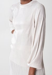 Lys creme bluse med vertikal ribbet tekstur, lange ærmer og høj krave. Stoffet fremstår blødt med en subtil glans.
