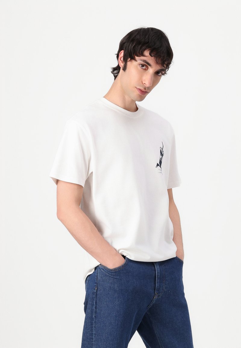 Camiseta de algodón blanca con cuello redondo, mangas cortas y un diseño gráfico negro en el lado izquierdo del pecho. Llevada con jeans azules.