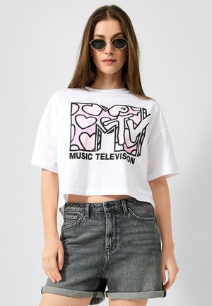 Młoda kobieta nosząca owalne okulary przeciwsłoneczne, białą, krótką koszulkę MTV Music Television z różowymi serduszkami oraz podwinięte ciemne krótkie spodenki dżinsowe na jednolitym tle.